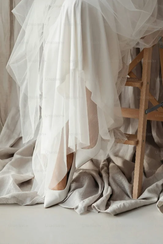 Jambes d'une femme assise sur un tabouret en robe de mariée avec des talons blancs